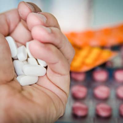 Outstretched hand holding several white pills. A colorful assortment of medication is slightly blurred in the background.